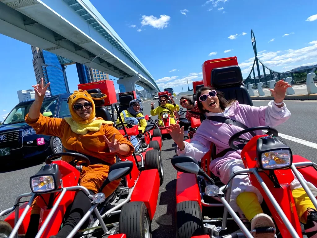IMG_0677 - StreetKart Group of friends in colorful onesies driving red go-karts on a sunny city road, waving at the camera under a large bridge.