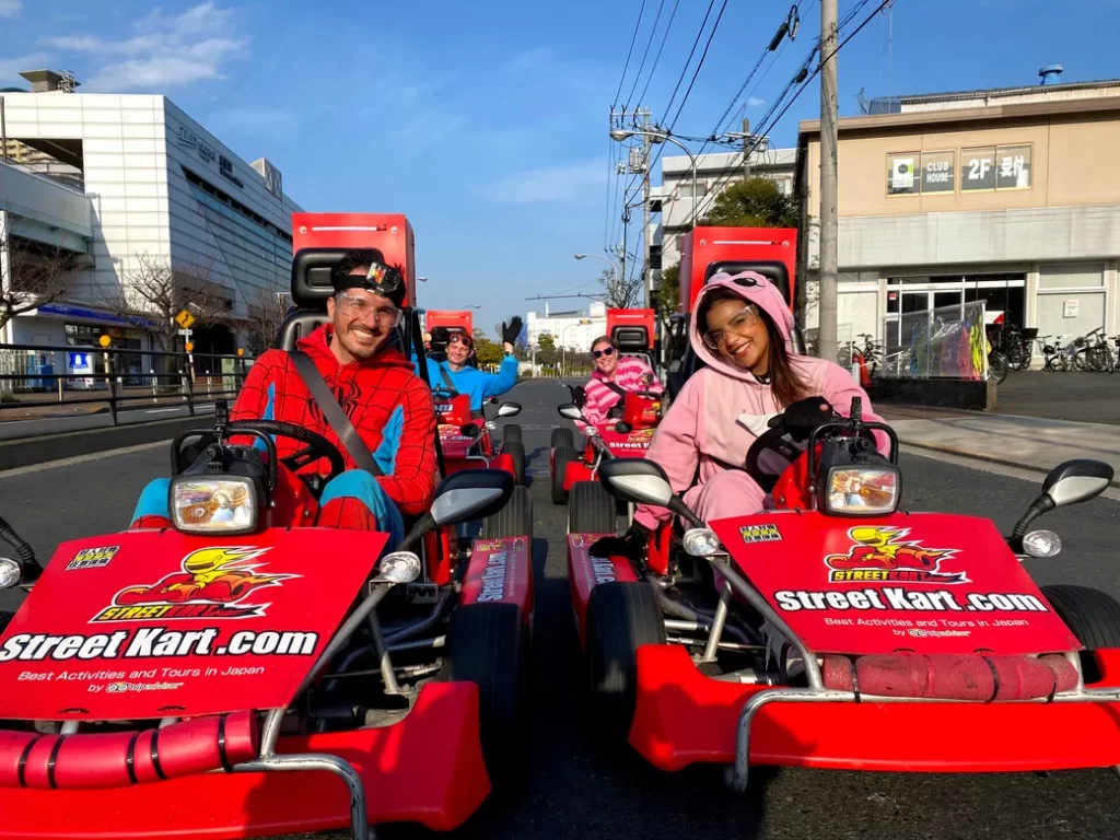 IMG_4101 - StreetKart People in red go-karts wearing colorful costumes smile and wave on a city street with Stadium branding on the carts.