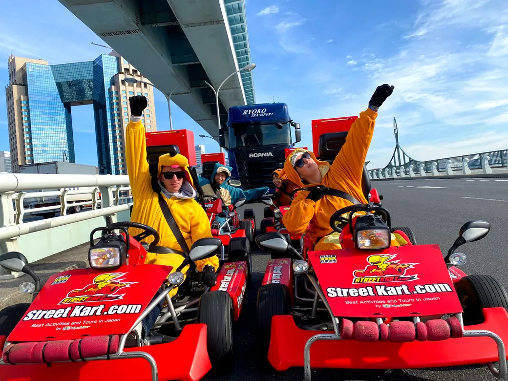 Group of people in yellow costumes driving red go-karts on a city bridge, waving at the camera