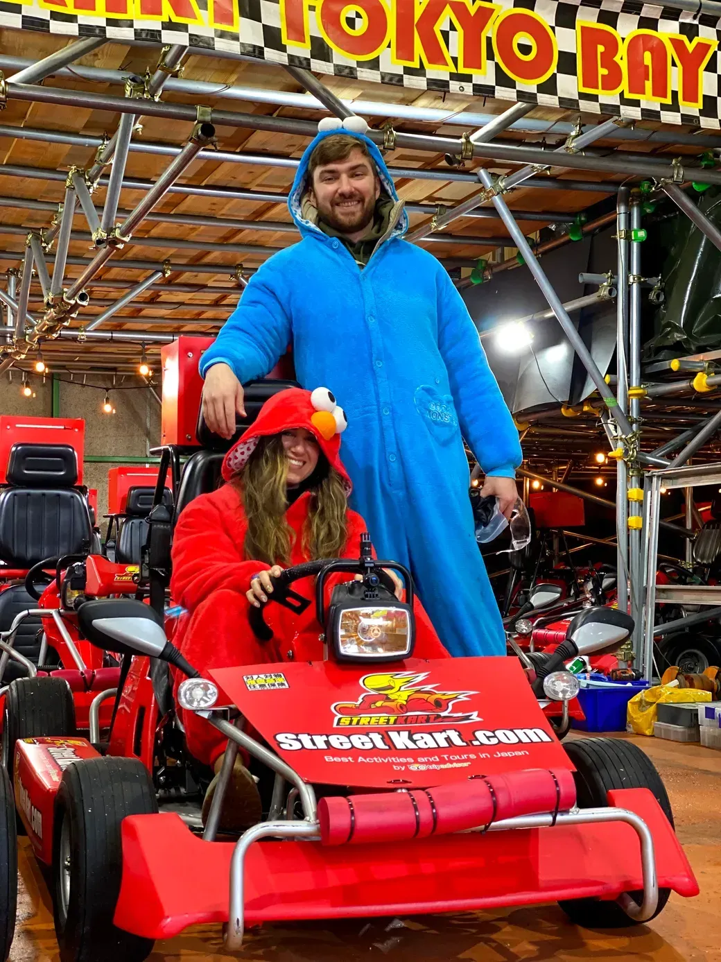 Two people in colorful costumes pose with a red go-kart indoors, a man in a blue onesie standing beside a woman in a red Elmo outfit, at a karting venue.